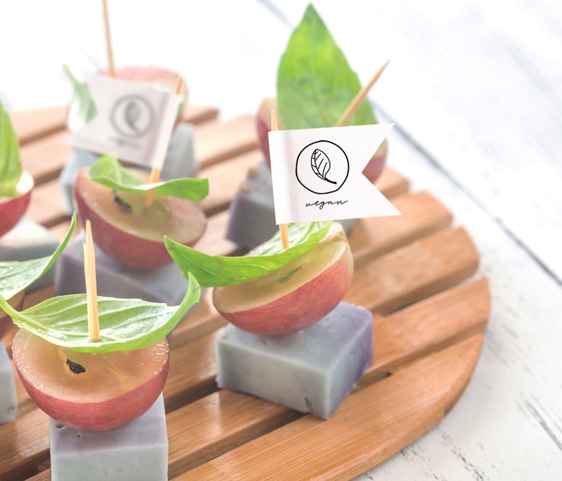 May include: A wooden platter with small purple and white squares topped with red grapes, basil leaves, and toothpicks with small flags that say "vegan".