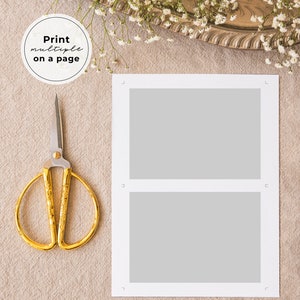 May include: A white card with two blank rectangular spaces for photos. The card is on a beige surface with a pair of gold scissors and a white flower arrangement.