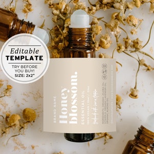 Pode incluir: Um rótulo bege com o texto "Honey blossom." e "ESSENTIAL OIL" em branco. O rótulo está preso a um frasco de vidro marrom com uma bola de rolamento branca. O frasco está rodeado de flores de camomila secas sobre um fundo branco.