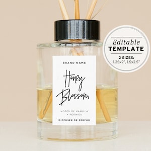 Op de afbeelding: Een heldere glazen diffuserfles met een zwarte dop en houten stokjes. Het etiket vermeldt "BRAND NAME", "Honey Blossom", "NOTES OF VANILLA + PEONIES" en "DIFFUSER DE PERFUM". Het etiket is wit met zwarte tekst. De fles staat op een lichtbruin oppervlak. Bewerkbare sjabloon 2 maten: 1,25x2", 1,5x2,5"