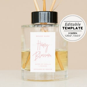Könnte beinhalten: Eine klare Glasdiffuserflasche mit schwarzem Deckel und Holzstäbchen. Das Etikett ist weiß mit dem Text "BRAND NAME", "Honey Blossom", "NOTES OF VANILLA + PEONIES", und "DIFFUSER DE PERFUM". Der Text "Honey Blossom" ist in rosa Schrift. Das Etikett enthält auch den Text "Editable TEMPLATE 2 SIZES: 1.25x2", 1.5x2.5"