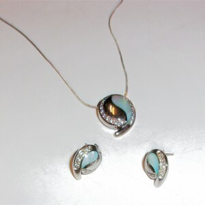 Puede incluir: Un collar de plata con un colgante de concha azul y blanca y pendientes a juego. El colgante está rodeado de pequeñas piedras transparentes.