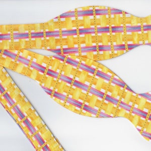 Puede incluir: Una pajarita amarilla con un patrón de cuadros rosa y morado. La pajarita está hecha de tela y tiene un acabado satinado.