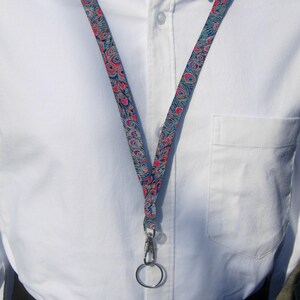 Puede incluir: Un cordón azul y rosa con estampado de plumas de pavo real, con un cierre giratorio plateado y un anillo plateado. El cordón está hecho de tela.