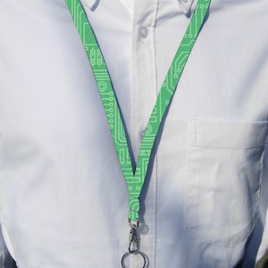 Puede incluir: Un cordón verde con un patrón de placa de circuito blanco. El cordón tiene un clip de metal plateado y un anillo en el extremo.