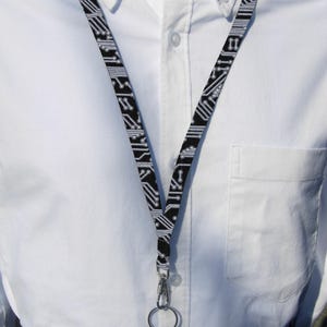 Pode incluir: Um cordão preto e branco com um padrão de placa de circuito. O cordão tem um clipe de metal prateado e um anel prateado.