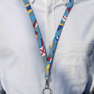 Könnte beinhalten: Ein blaues und gelbes Lanyard mit nautischem Thema. Das Lanyard hat Anker, Seile und Flaggen darauf gedruckt. Es hat einen silbernen Drehverschluss und einen silbernen Ring.
