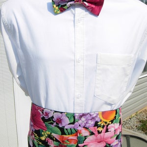 May include: A white dress shirt with a floral bow tie and a matching floral cummerbund. The bow tie and cummerbund feature a black background with pink, purple, and green flowers.