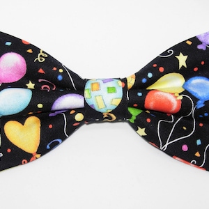 Puede incluir: Una pajarita negra con un estampado de globos de colores. Los globos son rojos, amarillos, azules, verdes y rosas. También hay confeti y serpentinas en la pajarita.