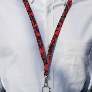 Może przedstawiać: Czerwono-czarny sznurek z wzorem i srebrnym karabińczykiem obrotowym. Sznurek nosi osoba w białej koszuli zapinanej na guziki.