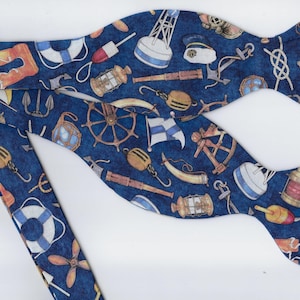 May include: A navy blue bow tie with a nautical pattern featuring anchors, life preservers, and ship wheels. The bow tie has a repeating pattern of maritime elements in shades of white, red, and gold.