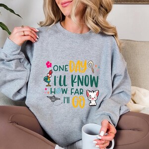 May include: Gray sweatshirt with a colorful graphic print featuring characters from the Disney movie Moana. The text on the shirt reads "One Day I'll Know How Far I'll Go."