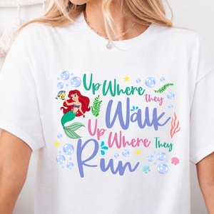 May include: White t-shirt featuring a colorful graphic of a mermaid with red hair and a green tail, accompanied by the text "Up Where They Walk, Up Where They Run." The design includes bubbles, seaweed, and seashells.