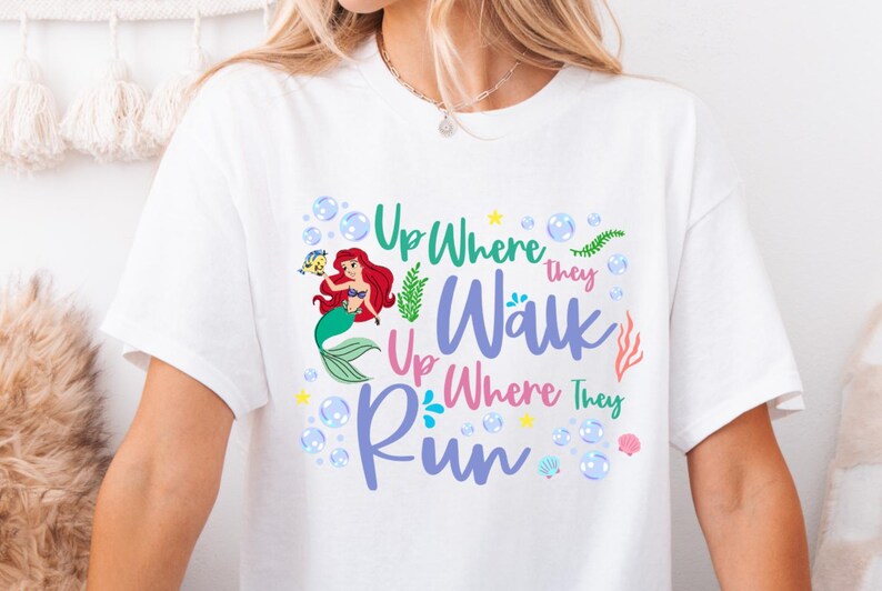 May include: White t-shirt featuring a colorful graphic of a mermaid with red hair and a green tail, accompanied by the text "Up Where They Walk, Up Where They Run." The design includes bubbles, seaweed, and seashells.