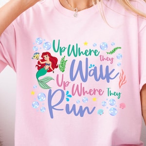May include: A light pink t-shirt featuring a colorful graphic of a mermaid with red hair and a green tail, surrounded by bubbles and aquatic plants. The text "Up Where They Walk, Up Where They Run" is displayed in various colors.