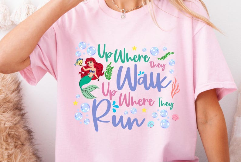 May include: A light pink t-shirt featuring a colorful graphic of a mermaid with red hair and a green tail, surrounded by bubbles and aquatic plants. The text "Up Where They Walk, Up Where They Run" is displayed in various colors.