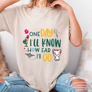 May include: A beige t-shirt with a graphic print of a rooster, a pig, a seashell, and a stick. The text on the shirt reads "One Day I'll Know How Far I'll Go."