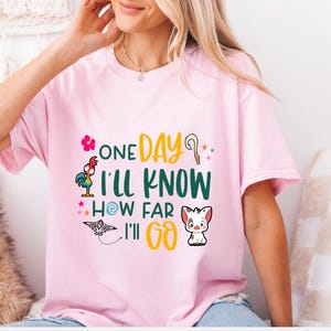 May include: Pink t-shirt with a graphic print of a rooster, a flower, a stick, a star, a swirl, a pig, and the text "One Day I'll Know How Far I'll Go."