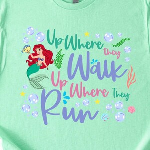 May include: Mint green t-shirt featuring a colorful graphic of a mermaid with red hair and a fish. The text reads "Up Where They Walk, Up Where They Run" in various colors, surrounded by bubbles, stars, and sea elements.