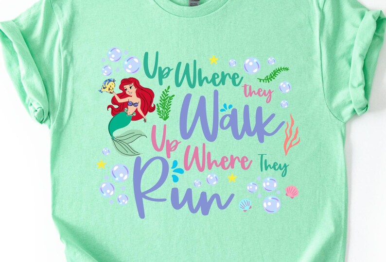 May include: Mint green t-shirt featuring a colorful graphic of a mermaid with red hair and a fish. The text reads "Up Where They Walk, Up Where They Run" in various colors, surrounded by bubbles, stars, and sea elements.