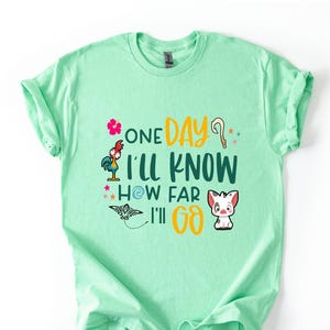 May include: A light green t-shirt with a graphic print of a rooster, a flower, a pig, and a seashell. The text on the shirt reads "One Day I'll Know How Far I'll Go."