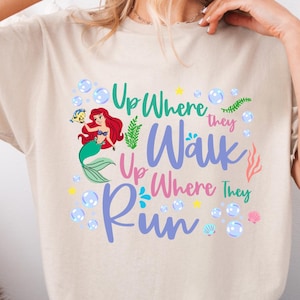 May include: Beige t-shirt featuring a colorful graphic of a mermaid with red hair and a green tail, surrounded by bubbles and the text "Up Where They Walk, Up Where They Run". The design includes sea elements.