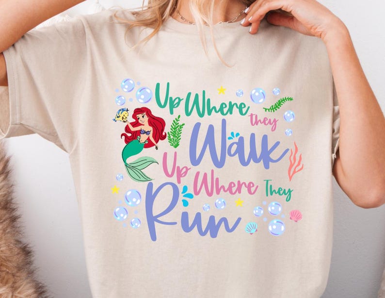 May include: Beige t-shirt featuring a colorful graphic of a mermaid with red hair and a green tail, surrounded by bubbles and the text "Up Where They Walk, Up Where They Run". The design includes sea elements.