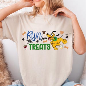 May include: Beige t-shirt featuring the phrase "Run for the Treats" in blue and green, with paw prints and bone graphics. A cartoon Pluto character is also featured in yellow and black.