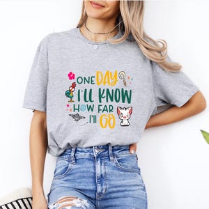 May include: A gray t-shirt with a graphic print of a rooster, a bird, and a pig. The text on the shirt reads "One Day I'll Know How Far I'll Go."