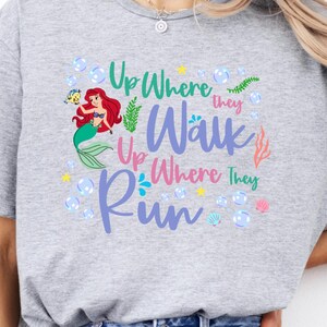 May include: A light gray t-shirt featuring a colorful graphic of a mermaid with red hair and a green tail, surrounded by bubbles and the text "Up Where They Walk, Up Where They Run."
