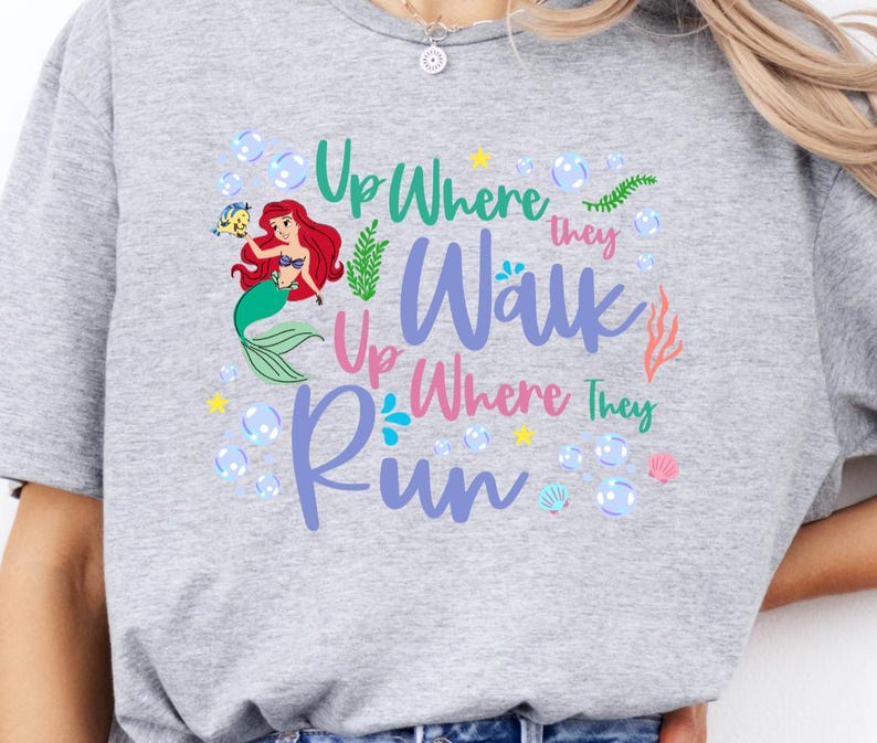 May include: A light gray t-shirt featuring a colorful graphic of a mermaid with red hair and a green tail, surrounded by bubbles and the text "Up Where They Walk, Up Where They Run."