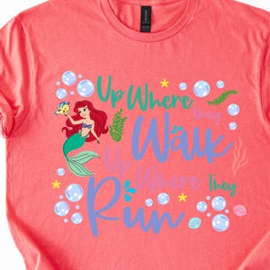 May include: Coral-colored t-shirt featuring a graphic of Ariel from The Little Mermaid, holding Flounder. The text reads "Up Where They Walk, Up Where They Run" in teal and purple, surrounded by bubbles and sea elements.