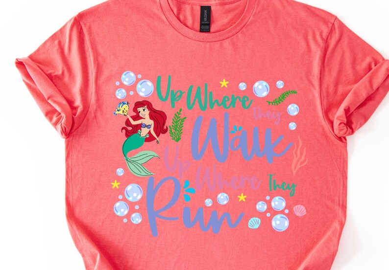 May include: Coral-colored t-shirt featuring a graphic of Ariel from The Little Mermaid, holding Flounder. The text reads "Up Where They Walk, Up Where They Run" in teal and purple, surrounded by bubbles and sea elements.