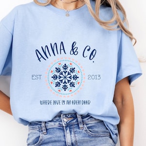 May include: Light blue t-shirt with the text "ANNA & CO." above a circular design featuring a snowflake and hearts. The text "EST 2013" and "WHERE LOVE IS AN OPEN DOOR" are also printed on the shirt.