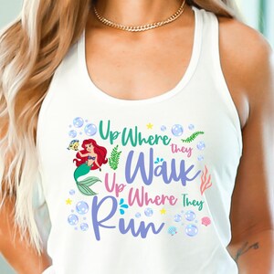 May include: White tank top featuring a colorful graphic of a mermaid with red hair and a green tail, accompanied by the text "Up Where They Walk, Up Where They Run." The design includes bubbles, seaweed, and stars.