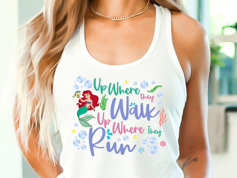 May include: White tank top featuring a colorful graphic of a mermaid with red hair and a green tail, accompanied by the text "Up Where They Walk, Up Where They Run." The design includes bubbles, seaweed, and stars.