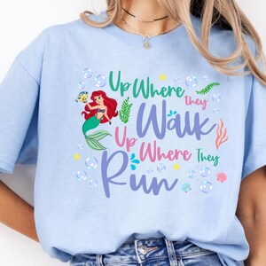 May include: Light blue t-shirt featuring a colorful graphic of a mermaid with red hair and a green tail, accompanied by the text "Up Where They Walk Up Where They Run". The design includes bubbles and aquatic elements.