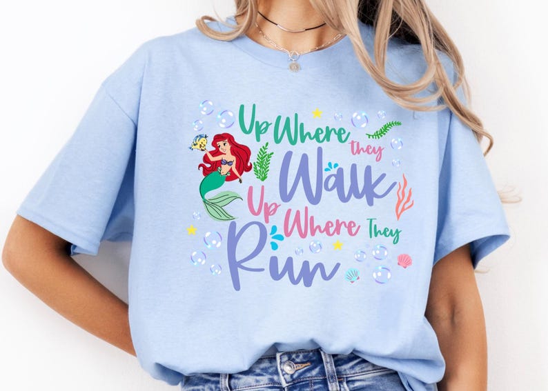 May include: Light blue t-shirt featuring a colorful graphic of a mermaid with red hair and a green tail, accompanied by the text "Up Where They Walk Up Where They Run". The design includes bubbles and aquatic elements.