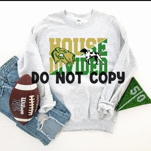 May include: A light gray sweatshirt with the words "HOUSE DIVIDED" in gold and green, featuring dog illustrations. A brown football and a green field marker are next to the sweatshirt. The words "DO NOT COPY" are also visible.