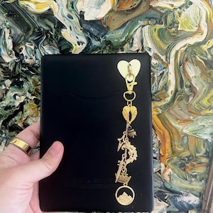 Puede incluir: Un lector electrónico negro con un dije dorado que presenta un corazón, alas, una estrella, una espada, una corona y una escena de montaña. El dije está sujeto al lector electrónico con un cierre dorado. El fondo es una pintura abstracta colorida.