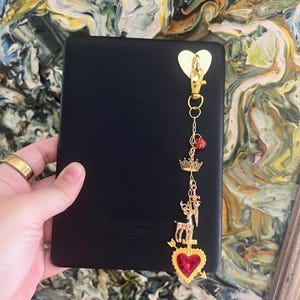 Puede incluir: Lector electrónico negro con un dije dorado. El dije presenta un cierre en forma de corazón, un colgante de corazón rojo, una corona, un ciervo y una cuenta roja. El lector electrónico se sostiene en una mano con un anillo dorado.