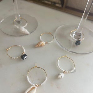 May include: A selection of gold-coloured wine glass charms. Each charm features a small seashell or decorative bead, strung on a white beaded hoop. The charms are designed to hang from the stem of a wine glass.
