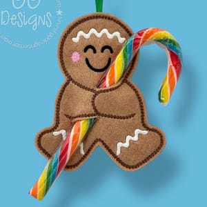 May include: A brown felt gingerbread man Christmas ornament with a smiling face, white icing details, and a green hanging loop. The gingerbread man is holding two rainbow-colored candy canes. The background is a solid light blue.