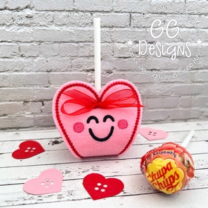 May include: A pink felt heart-shaped lollipop with a red ribbon and a smiling face. The lollipop is on a white stick and is surrounded by small red and pink hearts.