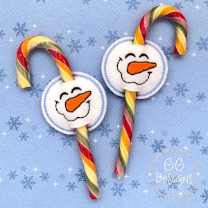 May include: Two candy canes with snowman faces attached. The candy canes are red, yellow, and green striped. The snowman faces are white with orange noses and black eyes.