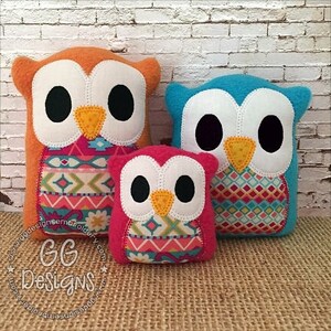 May include: Three plush owl toys in different sizes. The largest owl is orange with a pink, blue, and white geometric pattern. The middle owl is blue with a pink, blue, and white geometric pattern. The smallest owl is pink with a pink, blue, and white geometric pattern.