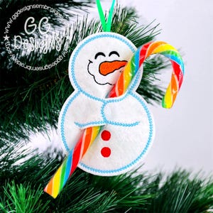 May include: A white snowman Christmas ornament with a smiling face, orange carrot nose, and blue stitched outline. The snowman holds a colorful rainbow candy cane. The ornament hangs from a green ribbon.