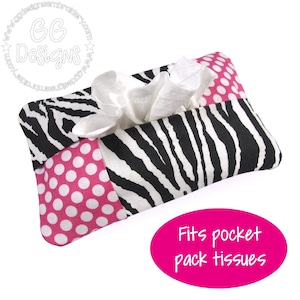 Puede incluir: Un porta-pañuelos rectangular con estampado de cebra y detalles de lunares rosas. El soporte dispensa pañuelos blancos y está diseñado para adaptarse a paquetes de pañuelos de bolsillo. El texto "Fits pocket pack tissues" se muestra en un óvalo rosa.