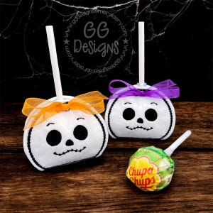 May include: Two white, pumpkin-shaped Halloween treats with black embroidered faces and stems, adorned with orange and purple ribbons. A Chupa Chups lollipop rests nearby. The background is dark with a spiderweb design.