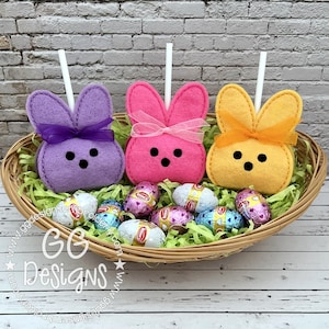 May include: Three felt Easter bunny pops in a wicker basket with green grass and chocolate eggs. The bunnies are purple, pink, and yellow with black eyes and a bow.
